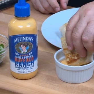 Hands drizzling Melinda's Ghost Pepper Ranch dressing from a bottle onto a tortilla wrap being prepared on a wooden surface