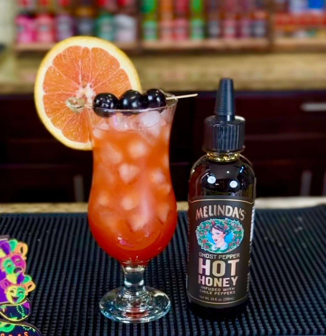 Orange cocktail with grapefruit slice and berries next to Melinda's Ghost Pepper Hot Honey bottle on bar mat
