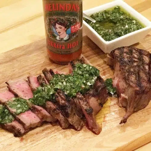 Sliced grilled steak topped with green chimichurri sauce on wooden cutting board, with hot sauce bottle and sauce bowl