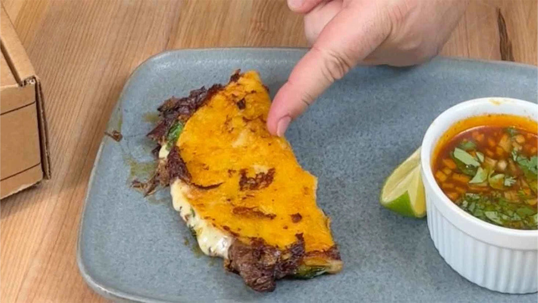 Hand pointing at birria quesadilla with melted cheese on blue plate, served with consommé dipping broth and lime wedge