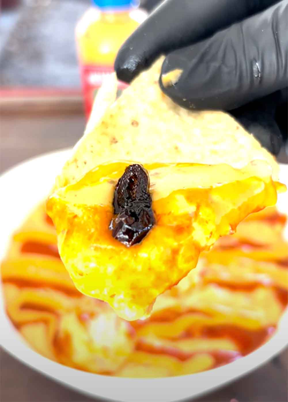 Person in black gloves holding tortilla chip with melted queso dip topped with dark chili pepper over bowl of layered...