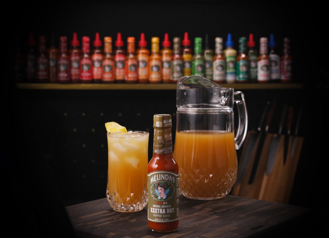 Mango tepache cocktail in glass with pineapple garnish, pitcher, and Melinda's XXXtra Hot sauce bottle on bar counter