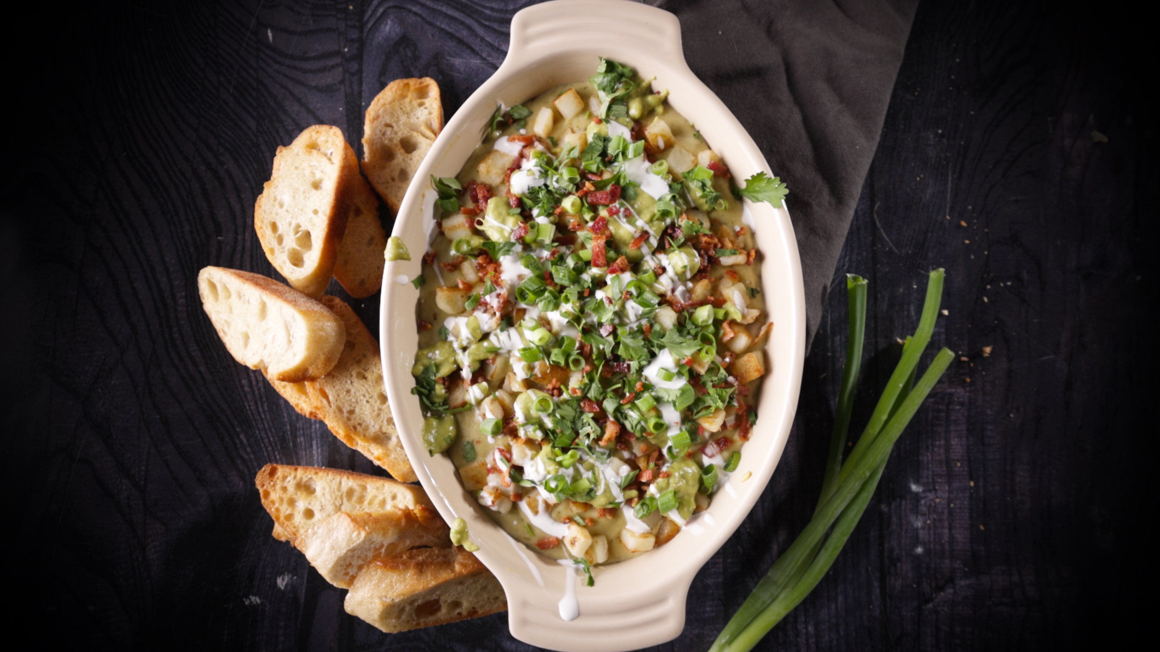 Creamy breakfast dip topped with herbs and bacon bits in white ceramic dish, served with sliced bread and green onions