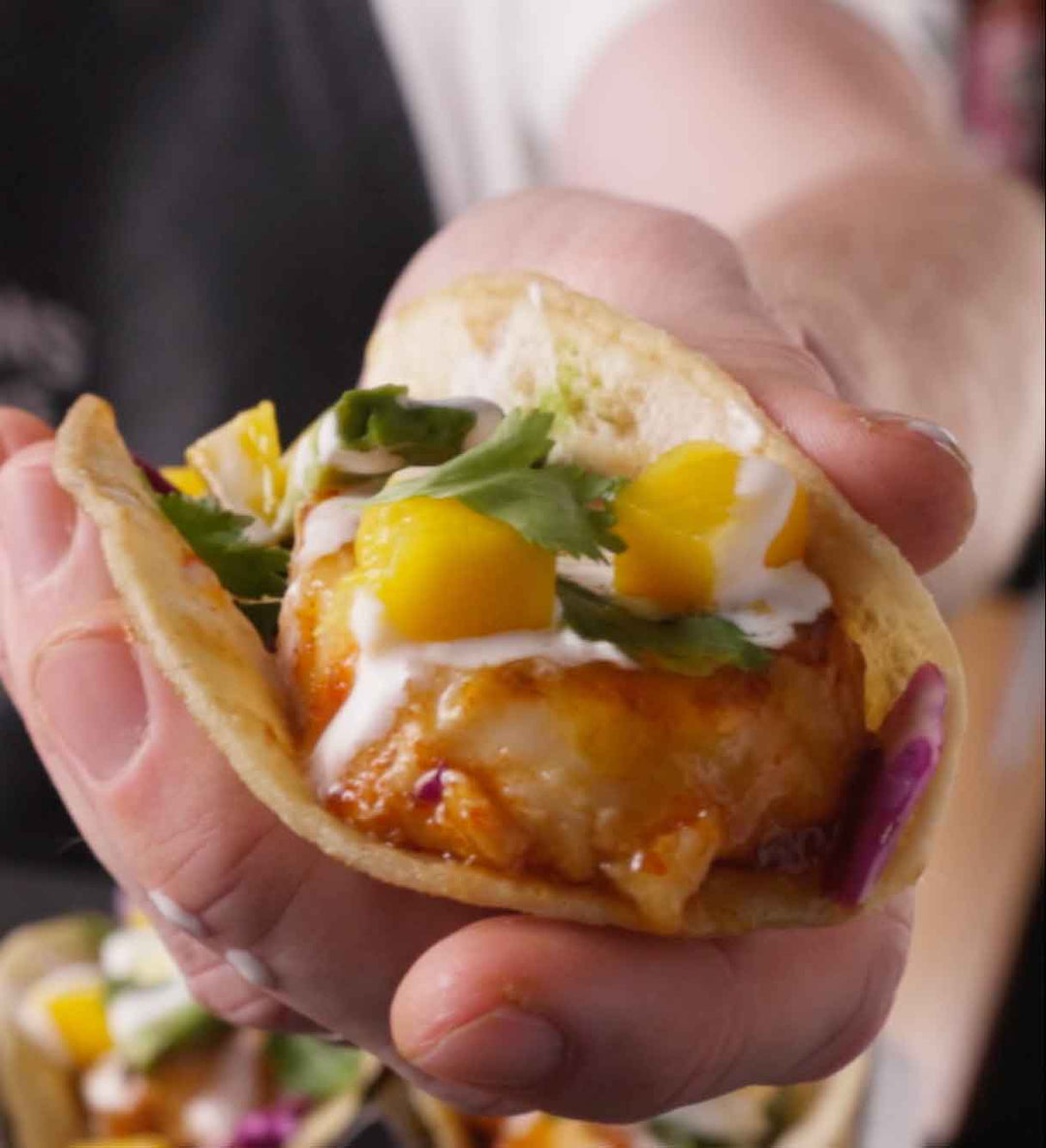 Hands holding a fish taco topped with mango, greens, and white sauce in a corn tortilla
