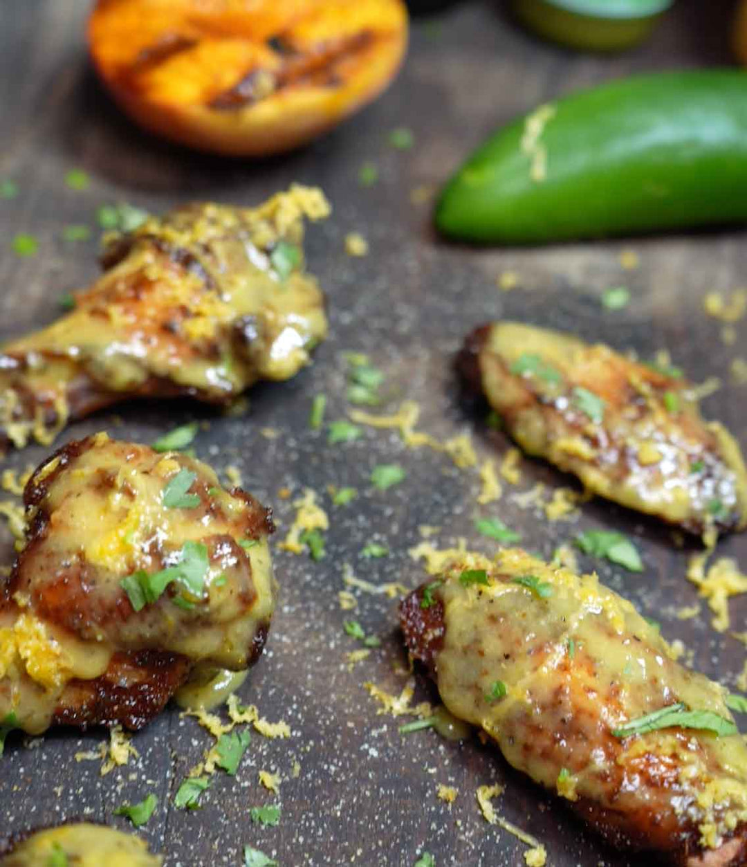 Grilled chicken drumsticks glazed with peach sauce, garnished with fresh herbs and lime zest on dark slate surface