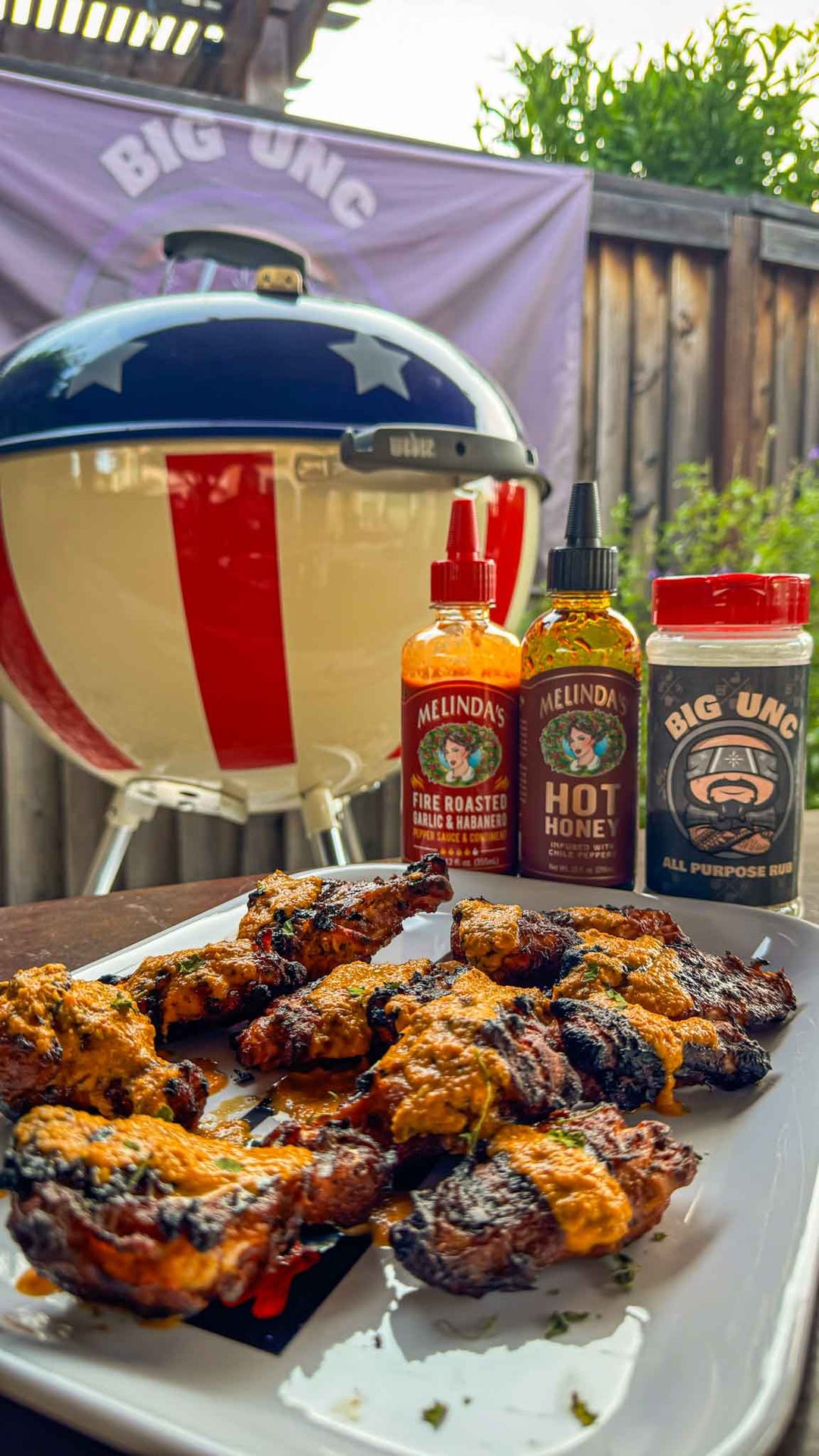 Grilled tikka masala wings on white plate with Melinda's hot sauces and Big Unc seasoning beside kettle grill outdoors