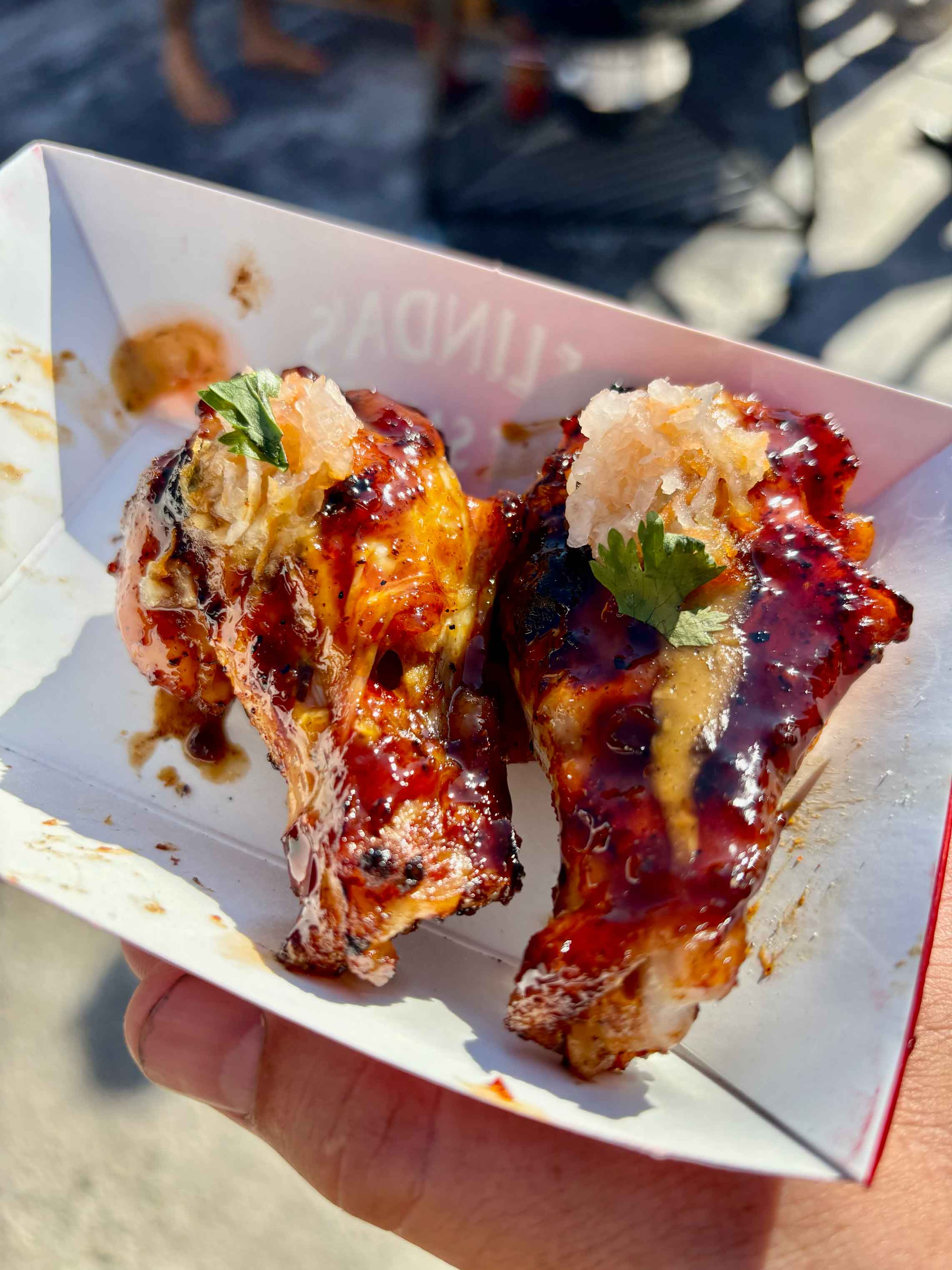 Two glazed chicken wings with dark sauce and fresh herbs served in a white paper container