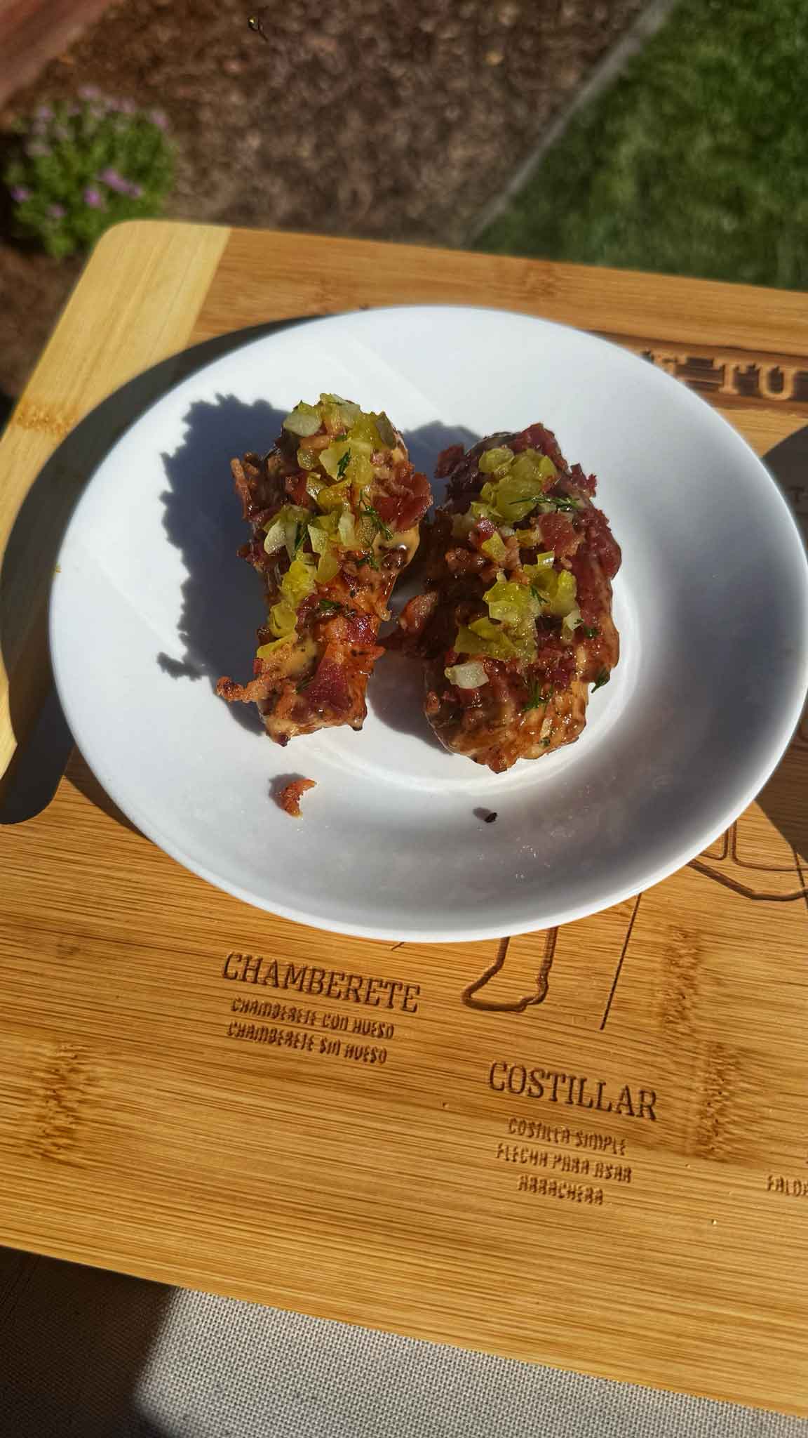 Two grilled chicken wings topped with green herbs on a white plate, placed on a wooden cutting board with Spanish text.