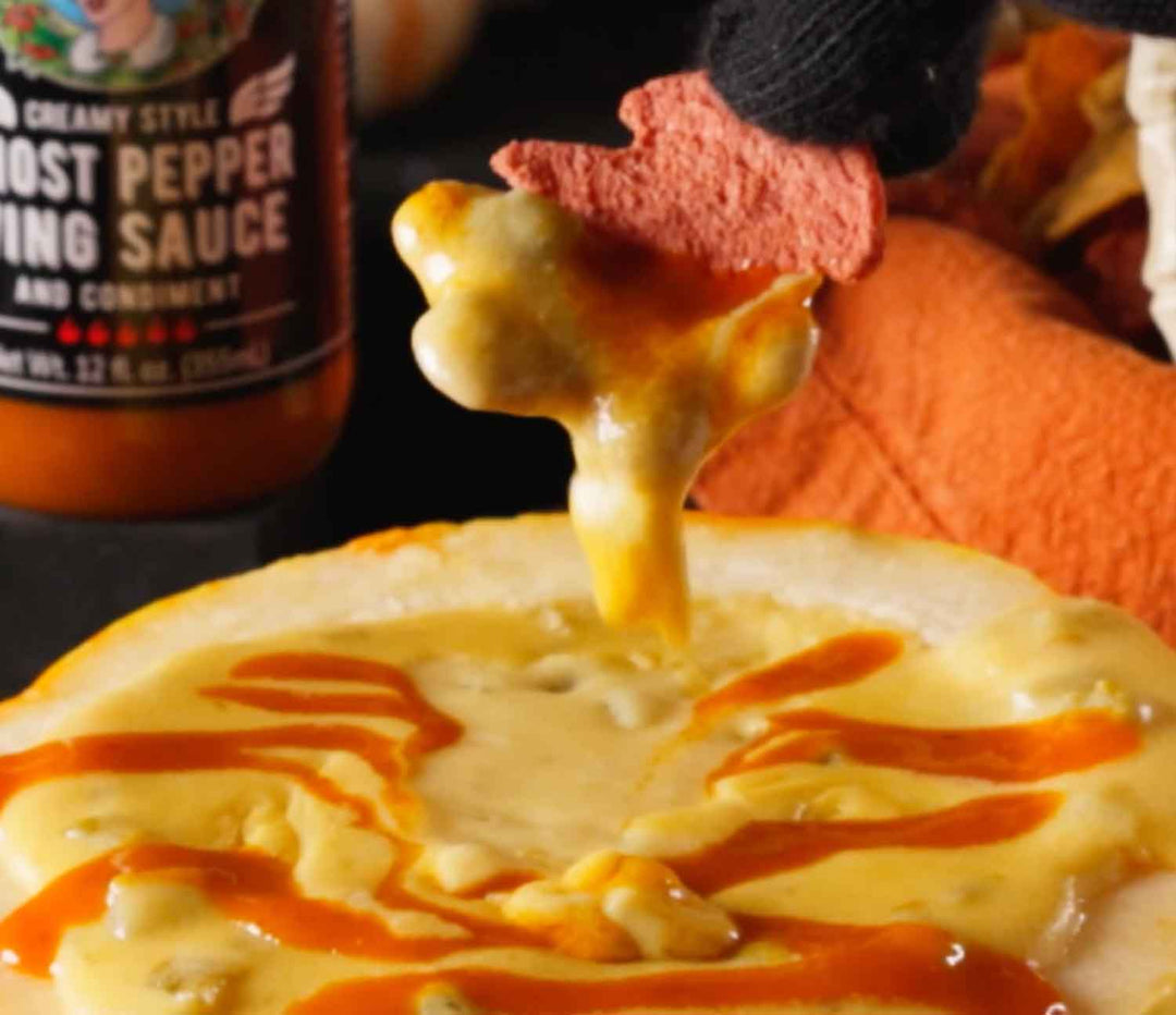 Melted cheese dripping from a spoon over bowl of queso with hot sauce swirls and ghost pepper bottle in background