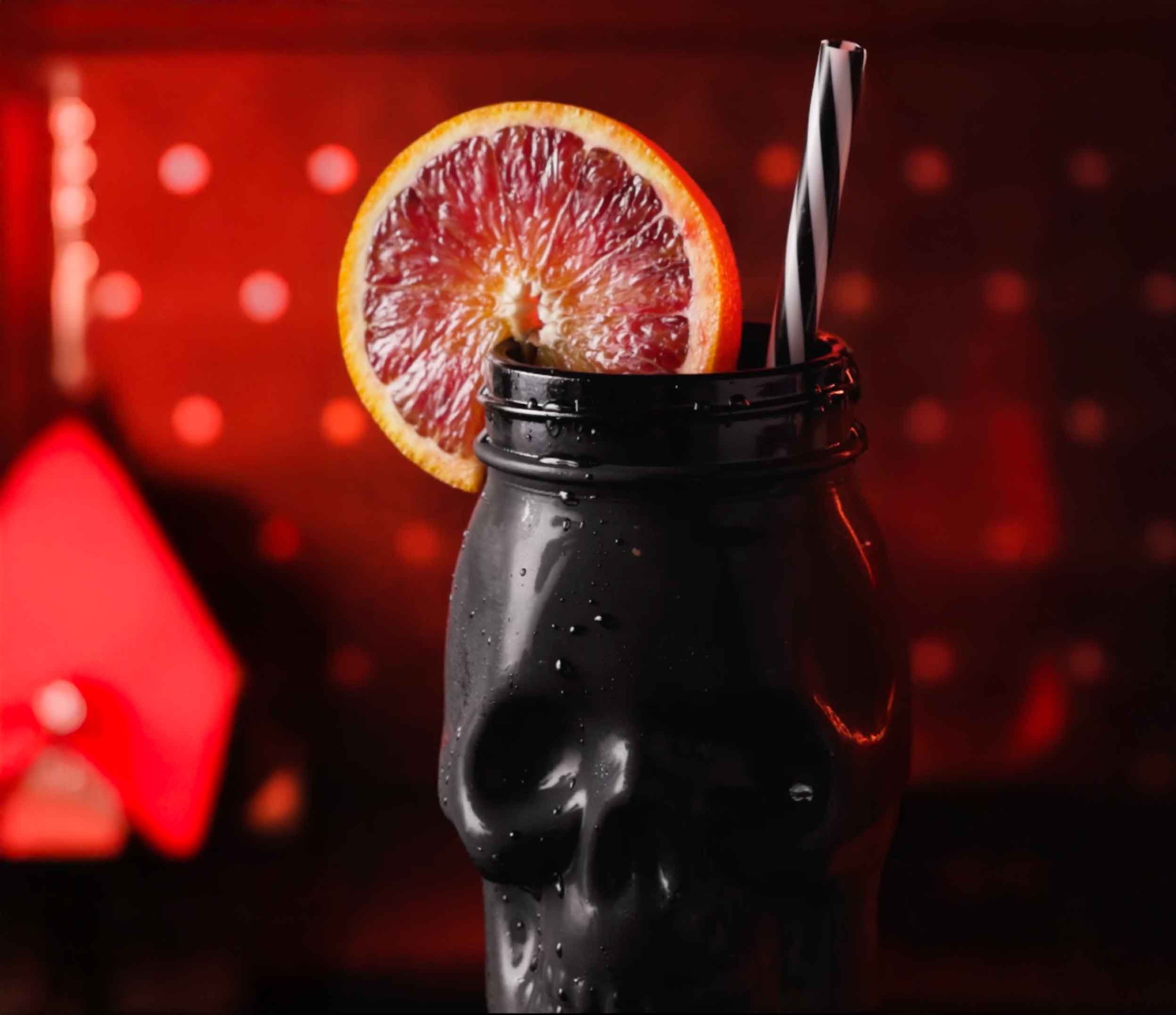 Dark glass bottle with grapefruit slice garnish and metal straw against red bokeh lights background