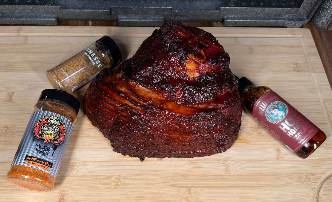 Glazed smoked ham with caramelized scoring on wooden cutting board, flanked by Cajun seasoning and hot honey sauce bottles