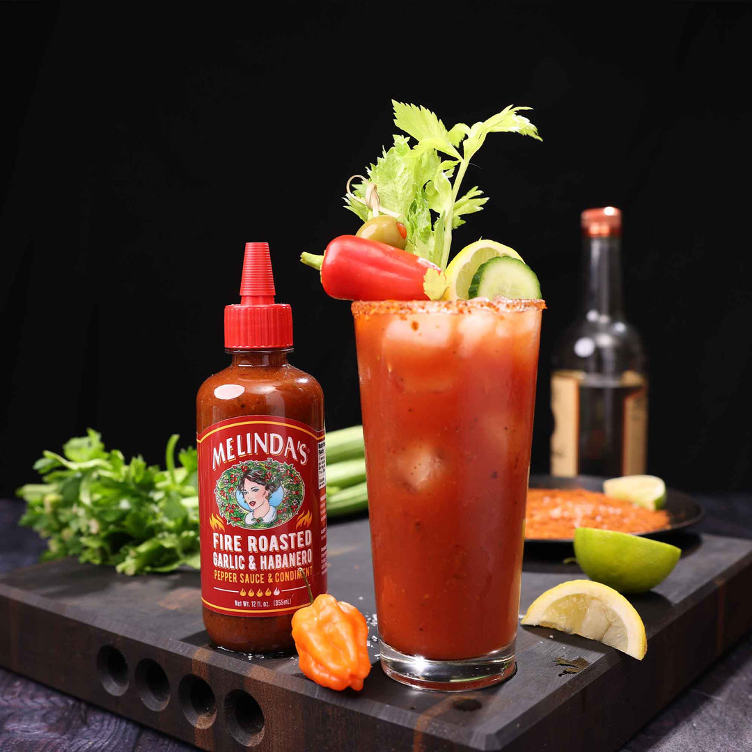 Bloody Mary cocktail garnished with celery and vegetables next to Melinda's Fire Roasted Garlic & Habanero hot sauce bottle