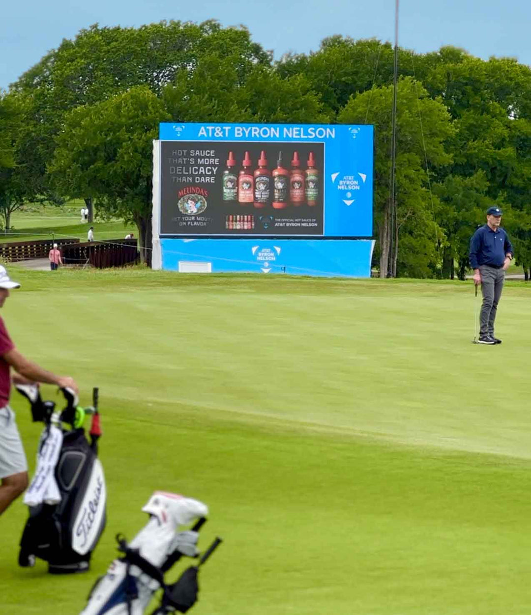 Golf course with AT&T Byron Nelson tournament signage featuring Melinda's hot sauce advertisement and golfer on green