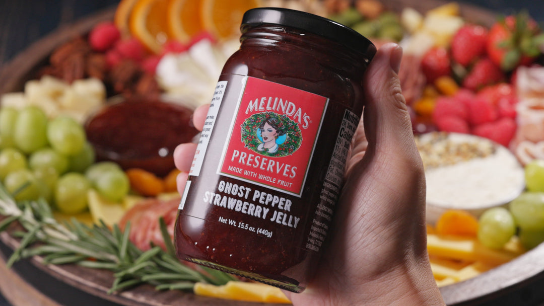 Hand holding jar of Melinda's Ghost Pepper Strawberry Jelly in front of colorful charcuterie board with fruits and cheese