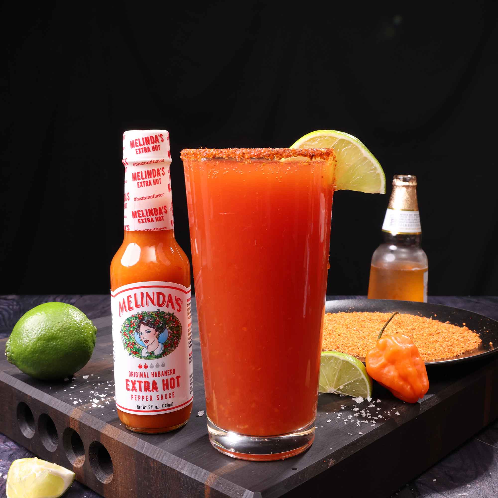 Spicy red michelada cocktail with lime garnish and salted rim, alongside Melinda's Extra Hot sauce bottle and fresh...