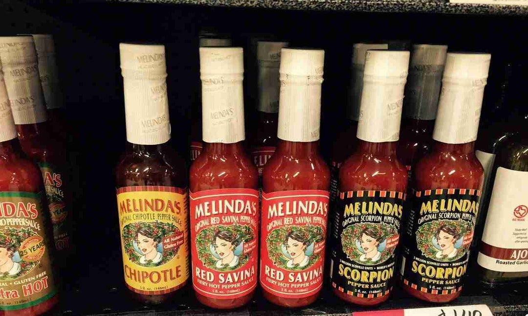Row of Melinda's hot sauce bottles on store shelf, including Chipotle, Red Savina, and Scorpion pepper varieties