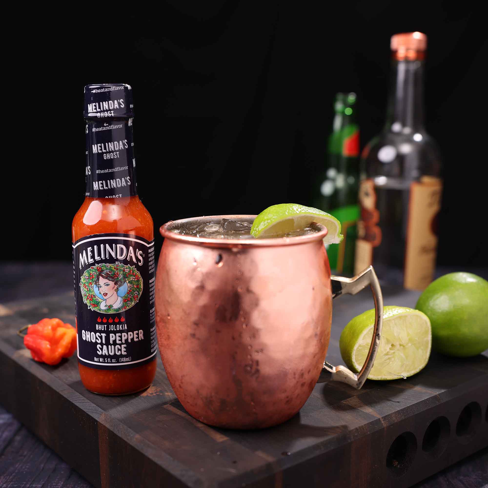 Copper Moscow mule mug with lime garnish next to Melinda's Ghost Pepper Sauce bottle on wooden board with lime wedges