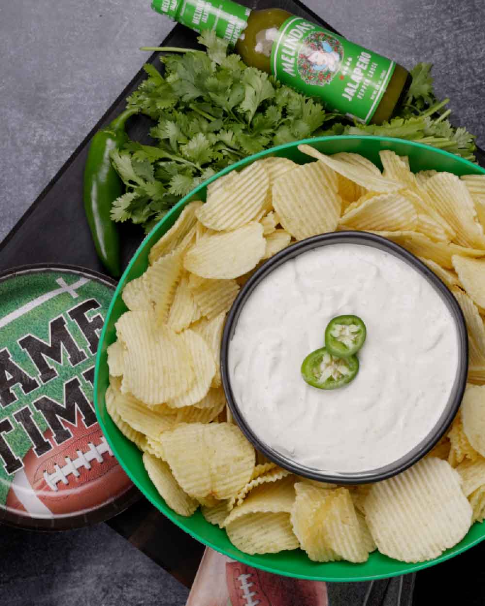Bowl of potato chips with white dip garnished with jalapeño slices, surrounded by football decorations and beer bottles