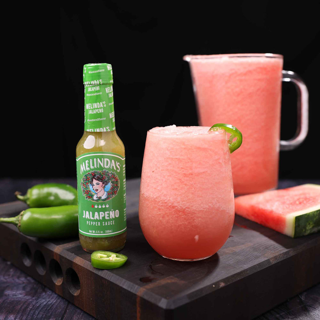 Pink watermelon frosé cocktail with jalapeño garnish next to Melinda's Jalapeño Pepper Sauce bottle and fresh ingredients