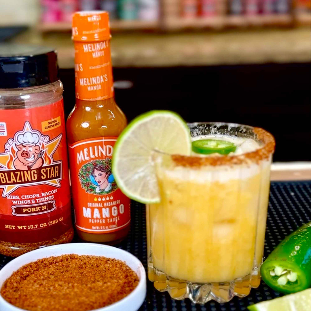 Mango margarita cocktail with lime garnish and jalapeño slice, alongside Melinda's mango hot sauce and Blazing Star seasoning