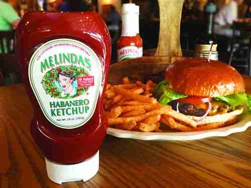 Melinda's Habanero Ketchup squeeze bottle next to a burger and sweet potato fries on a wooden table
