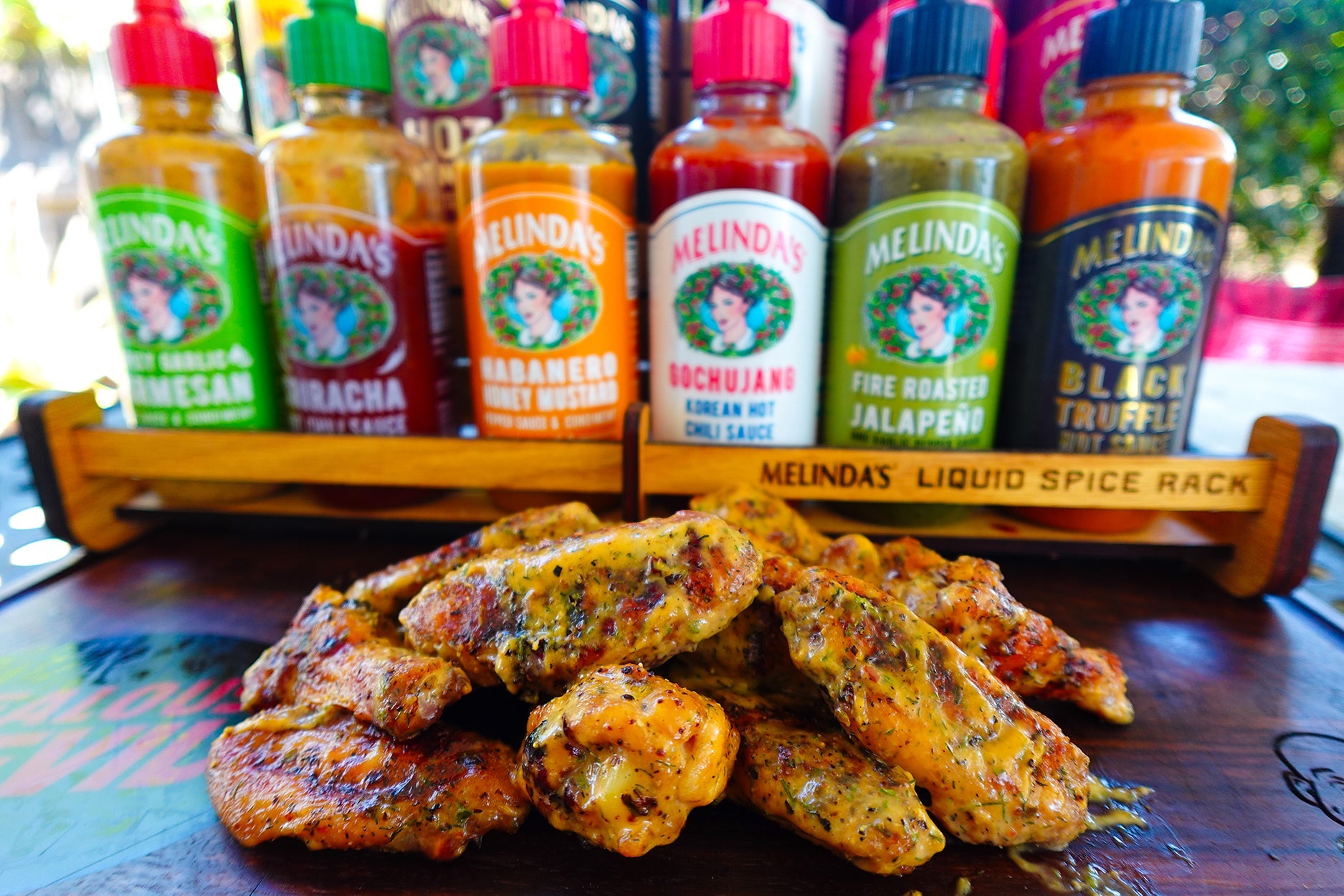Grilled chicken wings on a wooden table with a rack of six colorful Melinda's hot sauce bottles in the background.