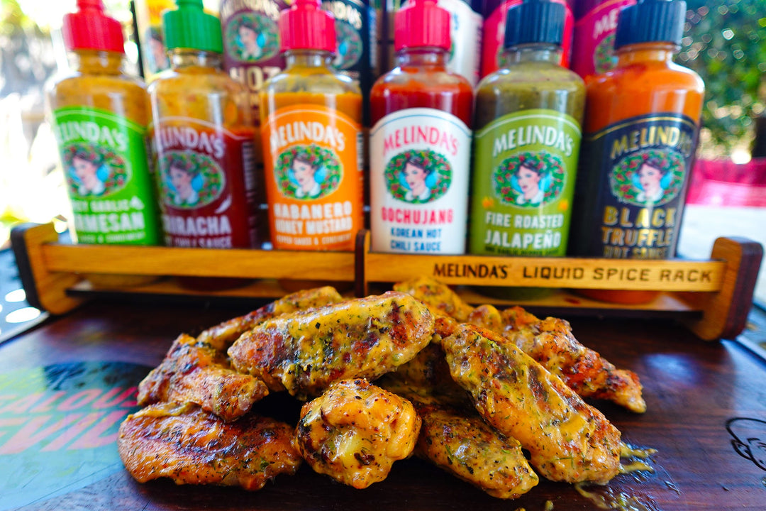 Grilled chicken wings on a wooden table with a rack of six colorful Melinda's hot sauce bottles in the background.