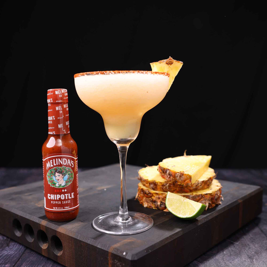 Pineapple chipotle margarita in coupe glass with spiced rim, garnished with pineapple wedge, beside hot sauce and lime
