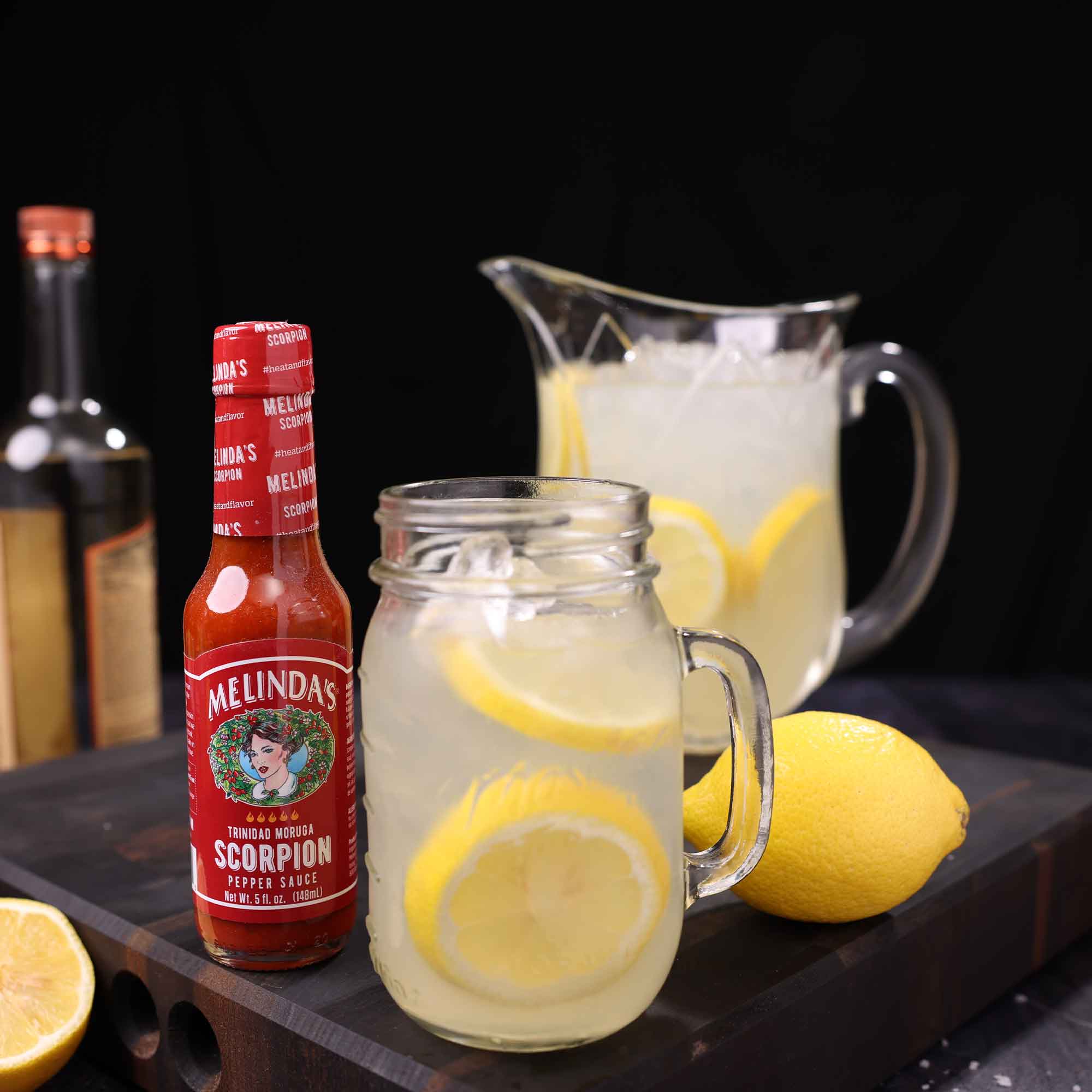 Mason jar mug of lemonade with lemon slices next to Melinda's Scorpion Pepper Sauce bottle, pitcher, and fresh lemons