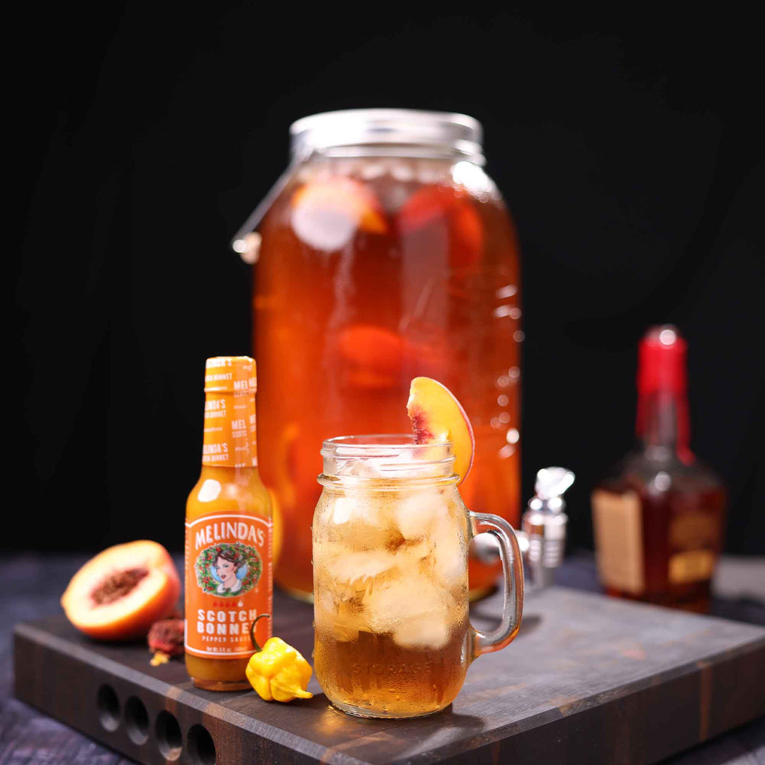 Bourbon peach tea cocktail in mason jar with Melinda's Scotch Bonnet sauce, fresh peaches, and whiskey bottle on wooden board