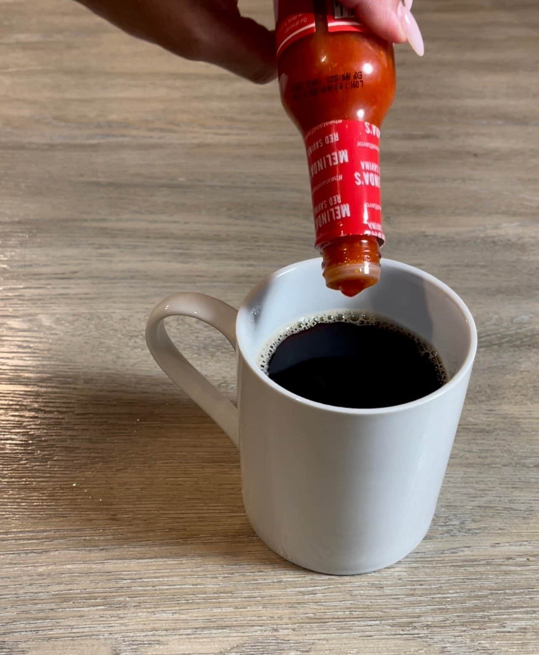 Hand squeezing hot sauce from red bottle into white coffee mug filled with black coffee on wooden surface