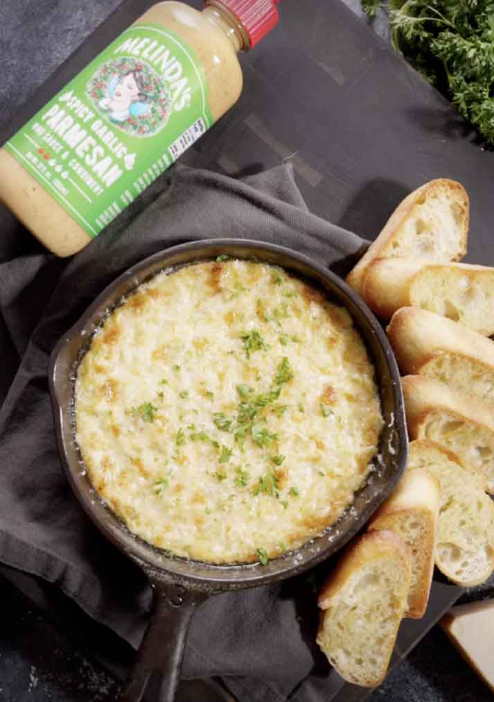 Hot artichoke dip in cast iron skillet with melted cheese and herbs, served with sliced bread and Melinda's sauce bottle