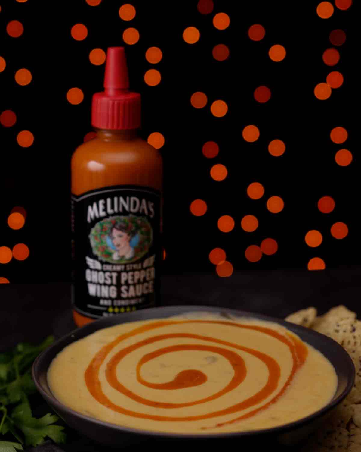 Bowl of creamy white queso with orange hot sauce swirl next to Melinda's Ghost Pepper Wing Sauce bottle on black background
