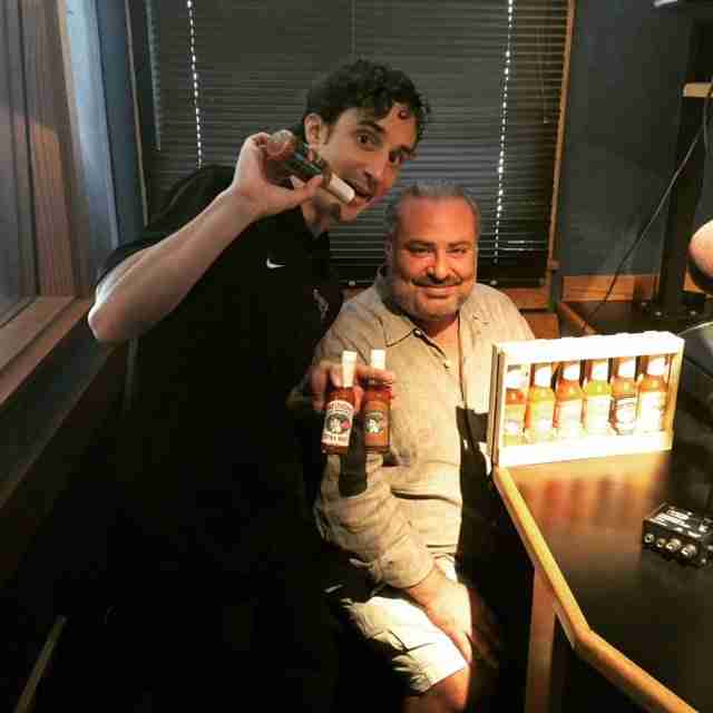 Two men in a radio studio, one standing with microphone, one seated, with bottles of Melinda's hot sauce displayed on desk