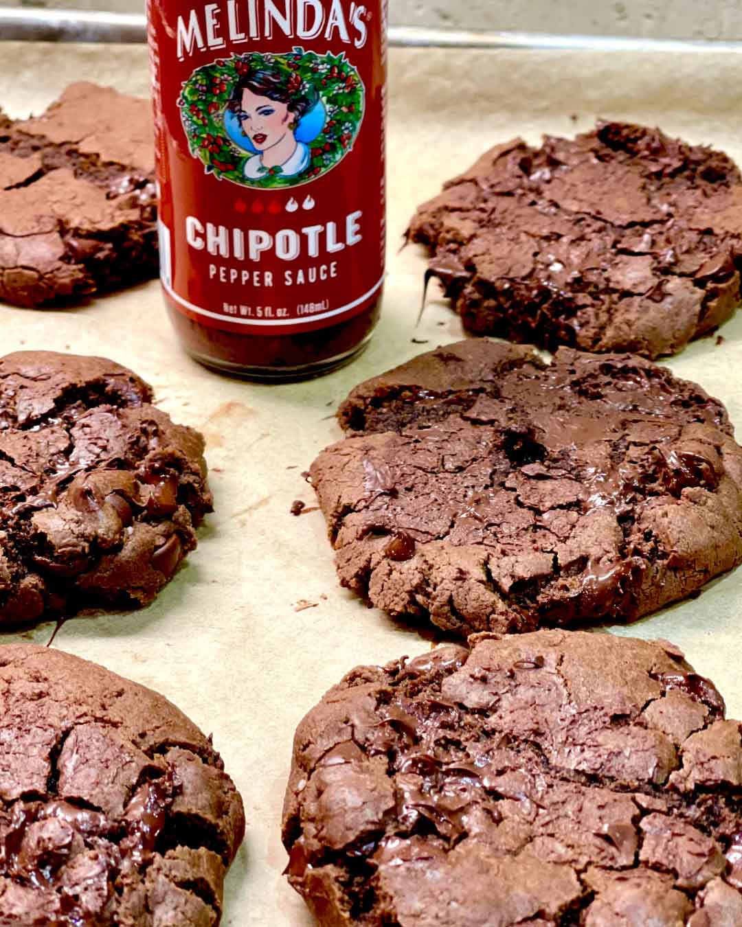 Double chocolate cookies with chocolate chips on parchment paper next to a bottle of Melinda's Chipotle Pepper Sauce