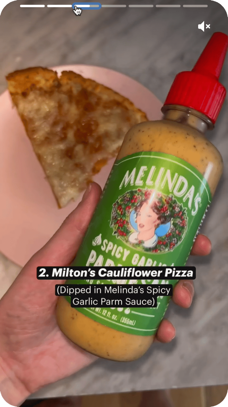 Hand holding Melinda's Spicy Garlic Parm Sauce bottle next to cauliflower pizza slice on pink plate