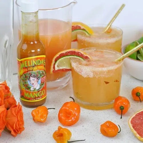 Spicy paloma cocktails garnished with grapefruit and lime slices, served with Melinda's mango hot sauce and orange peppers