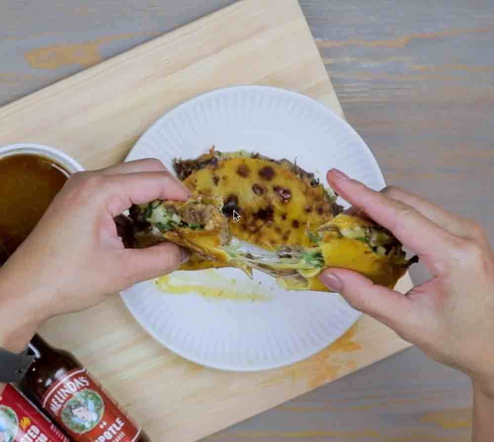 Hands holding a crispy birria tacodilla filled with melted cheese, meat, and cilantro on a white plate