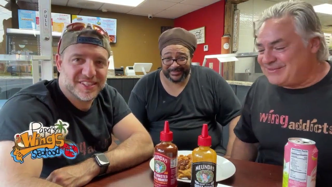 Three men sitting at restaurant table with two bottles of Melinda's hot sauce and food, smiling at camera